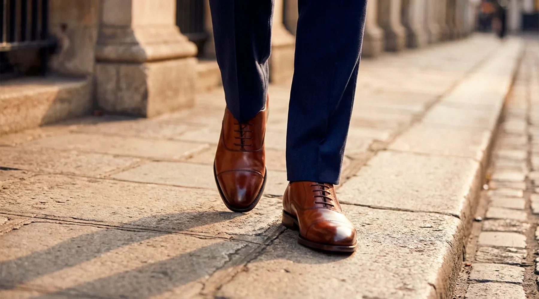 Primer plano de zapatos Oxford de piel marrón oscuro bien lustrados, combinados con el bajo de un pantalón de traje azul marino a medida. Detalle de estilo y coherencia en vestuario profesional.